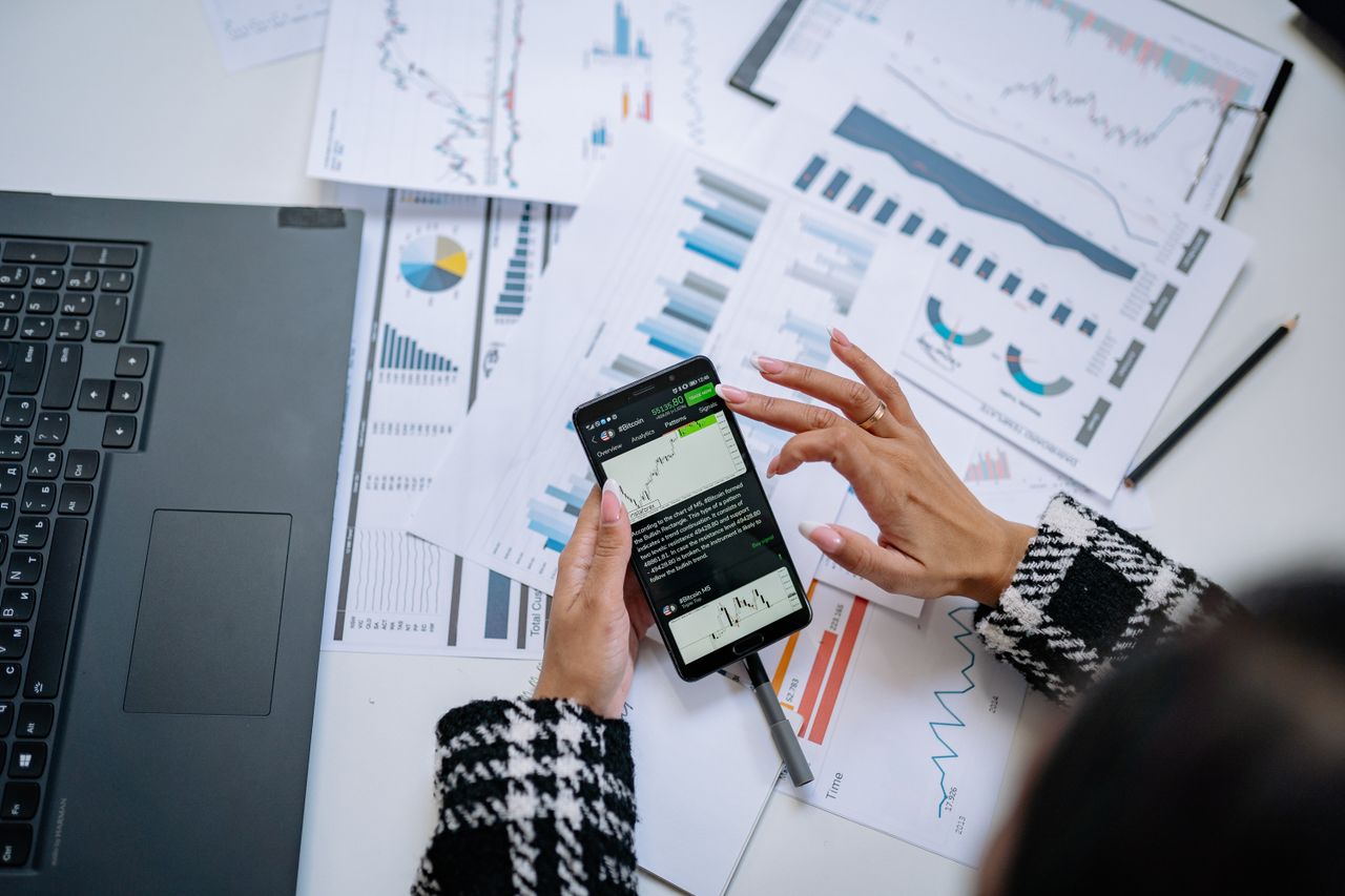 Femme d'affaires examinant des graphiques financiers sur smartphone, accompagnée d'un ordinateur portable et de documents.